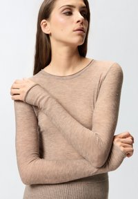 Žena s dugom smešnom kosom koja nosi prilagođenu, svetlosmeđu majicu dugih rukava sa suptilnim sjajem, pozira s prekriženim rukama i gleda u stranu.