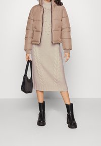 Giacca puffer beige sopra un abito midi a maglia cable color crema. Stivali neri con piattaforma e una borsa tote nera completano l'outfit. Materiali con texture evidenti.