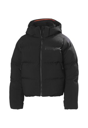 Niño con casco de esquí negro, gafas tintadas de rojo, chaqueta acolchada negra con logotipo, guantes a juego y pantalones negros de pie contra un fondo liso.