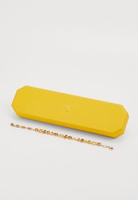 Gouden armband met afwisselende gele en amberkleurige edelstenen naast een gestructureerde gele octagonale sieradendoos op een witte ondergrond.