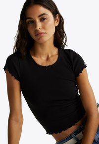 Czarny prążkowany top crop z krótkimi, falbaniaste rękawami, falbaniastym wykończeniem dołu i małym kokardkowym detalem przy dekolcie, zestawiony z niebieskimi dżinsami.