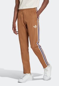 Pantalones deportivos marrones hechos de una tela suave, con un logo blanco de Adidas y rayas verticales moradas en los lados. Cintura elástica y bolsillos.