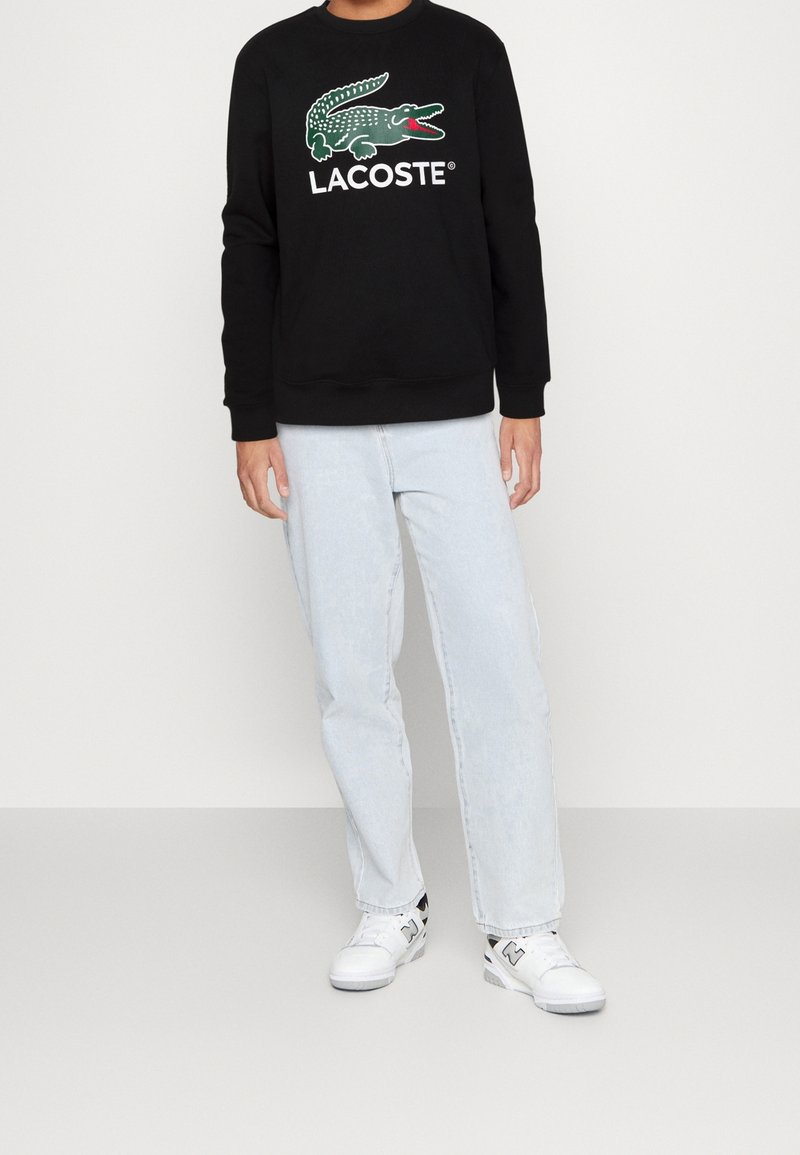 Personne portant un sweat-shirt noir Lacoste avec un logo crocodile vert, un jean bleu clair et des baskets blanches, debout devant un fond uni.