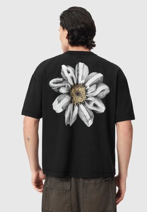 Uomo con maglietta nera con grande motivo di margherita bianca e gialla sul retro, abbinata a pantaloncini cargo scuri.