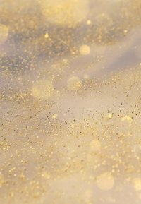 Złote drobinki brokatu rozsiane na miękkim, jasnym tle, tworząc migotliwy efekt z różnymi rozmiarami i teksturami brokatu.