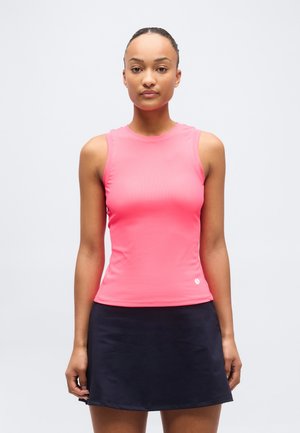 Žena oblečená v bezrukávovom jasne ružovom topu a čiernej sukni, stojaca čelom dopredu na neutrálno svetlom pozadí.