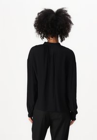 Blusa nera a maniche lunghe con colletto alto, vestibilità comoda e dettagli arricciati sul retro. Tessuto morbido con una superficie liscia.
