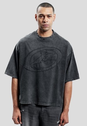 Homme aux cheveux courts et boucles d'oreilles portant un t-shirt noir délavé et ample avec un grand logo ovale et un jean foncé devant un fond gris.