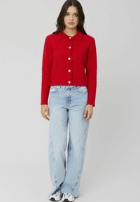 Cardigan rouge en maille côtelée avec un bord festonné, des fermetures à boutons et un col chemise, associé à un jean bleu clair à jambes larges et des baskets.