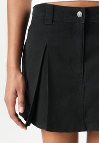 Svart denim plissert skjørt med knappelukking, synlige sømmer og en lett utvidet nederkant. Har en avslappet passform og glatt tekstur.