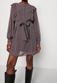 Langärmliges, gemustertes Kleid in dunklen Farbtönen mit Rüschendetails, einer knopfverzierten Ausschnittöffnung und einem gebundenen Gürtel in der Taille, kombiniert mit kniehohen schwarzen Stiefeln.