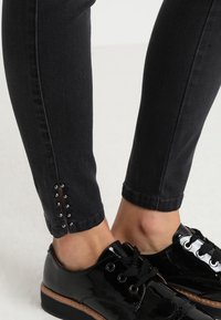 Černé úzké džíny s těsným střihem, zdobené rozparkem u kotníku a stříbrnými nýty. Měly by být kombinovány s lesklými černými platformovými botami.