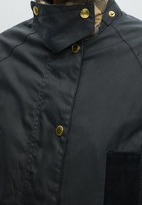 Κοντινή λήψη μαύρου παλτού με χρυσά κουμπώματα και εσωτερικό γιακά με καρό μοτίβο.