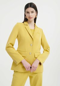 Blazer giallo su misura con due bottoni, revers a punta e tasche frontali. Il tessuto è liscio con bottoni lucidi.