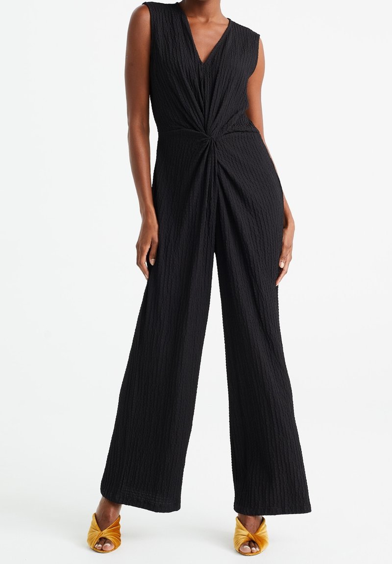 Czarny, teksturowany kombinezon z głębokim dekoltem w kształcie litery V i skręconym detalem w talii. Szerokie nogawki. W połączeniu z żółtymi sandałami na obcasie z paskami.