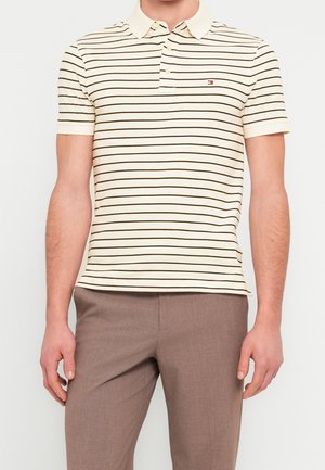Mand iført cremet kortærmet poloshirt med tynde sorte vandrette striber og brune bukser mod en ensfarvet baggrund.