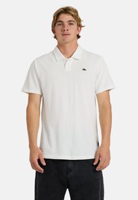 Νεαρός άνδρας με ξανθά μαλλιά, φορώντας άσπρο πόλο πουκάμισο και μαύρο παντελόνι, στέκεται μπροστά σε ένα απλό λευκό φόντο.