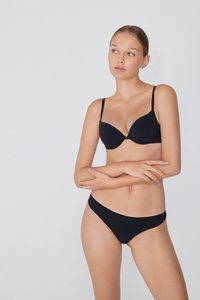 Mladá žena se světlou pletí a staženými vlasy, oblečená v černé podprsence a odpovídajícím černém spodním prádle, stojí s překříženýma rukama na jednobarevném pozadí.