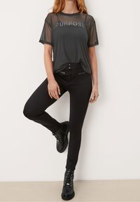 Sort mesh t-shirt med teksten "PURPOSE", parret med sorte slim-fit bukser med frontlynlåse og quilted detaljer, båret med sorte støvler.
