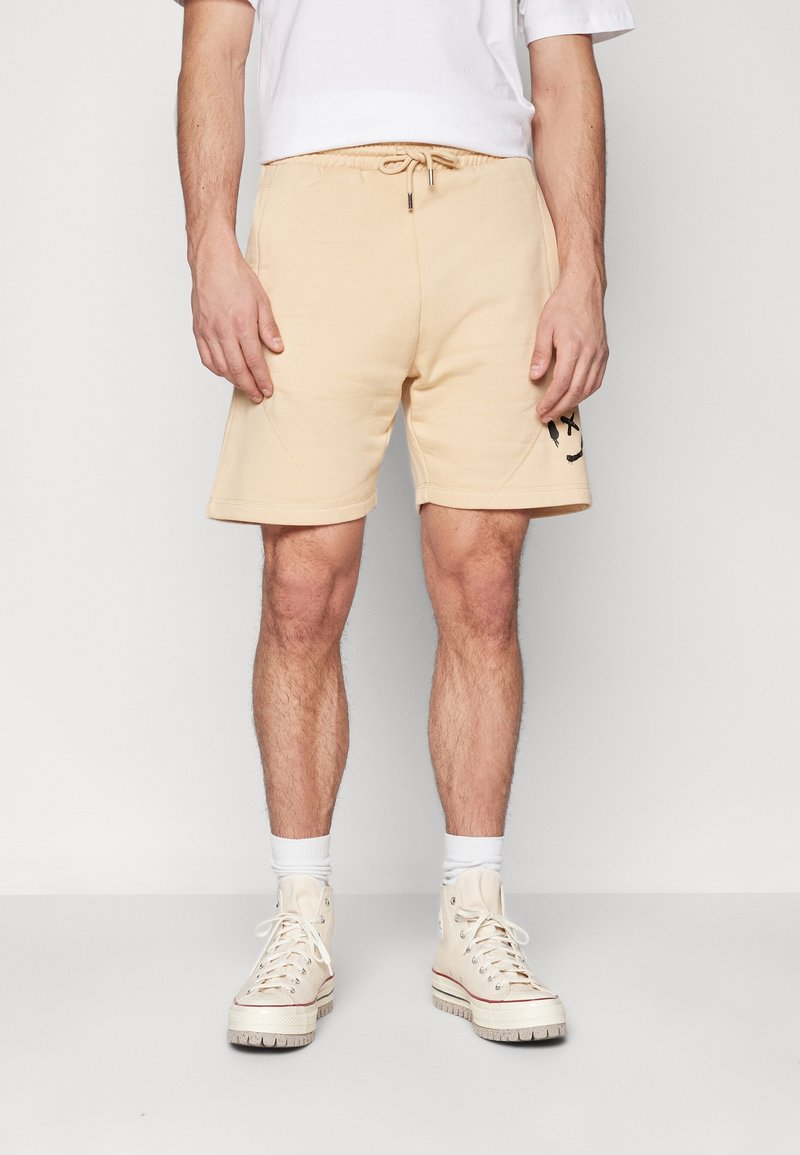 Shorts en coton beige avec une ceinture élastique et un cordon de serrage. Présente un motif de smiley noir sur la cuisse gauche.
