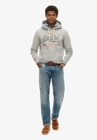 Grå hættetrøje med et trykt logo, blå jeans og brune læderstøvler. Hættetrøjen har snoretræk og en frontlomme.
