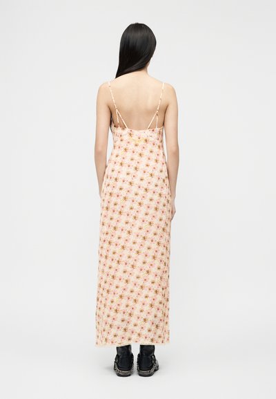 Γυναίκα με μακριά σκούρα μαλλιά, που φοράει αμάνικο, ροζ μακρύ φόρεμα με λουλούδια και μαύρες μπότες, στέκεται με την πλάτη στραμμένη προς τα πίσω σε απλό φόντο.