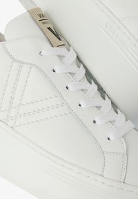 Weiße Ledersneaker mit flachen weißen Schnürsenkeln, detaillierter Naht und einer strukturierten Gummisohle. Das Etikett auf der Zunge zeigt die Initialen der Marke.