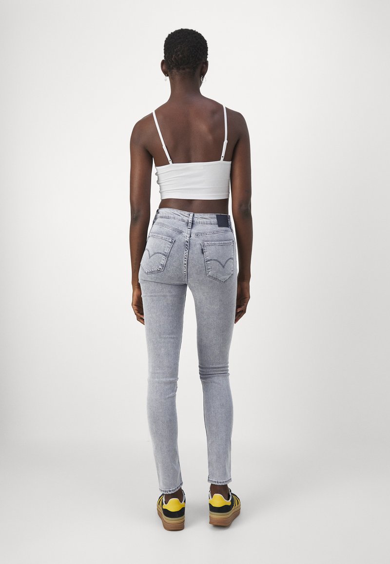 Schlanke, hellblaue Jeans mit hoher Taille, fünf Taschen und einem Logo-Patch auf der Rückseite. Kombiniert mit einem weißen Crop-Top und Sneakers.