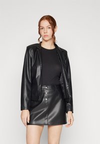 Blazer en simili cuir noir avec un col cranté, associé à un haut noir ajusté et une mini-jupe en simili cuir noir taille haute, dotée de boutons-pression.