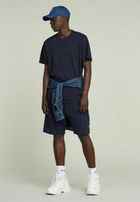 T-shirt navy con colletto rotondo, abbinata a pantaloni corti navy e una giacca di jeans legata in vita. Sneakers bianche e un cappellino di jeans.