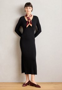 Robe noire en tricot côtelé avec décolleté en V, manches longues et fente sur le côté. Associée à un foulard en soie maroon et doré à motifs et des chaussures à talons carrés bordeaux.