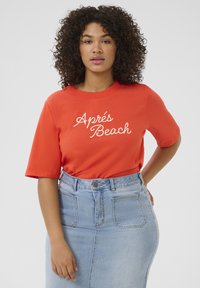 T-shirt en coton orange vif à manches courtes, avec un texte en cursive blanche "Après Beach". Associé à une jupe en denim bleu clair.