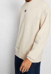 Person wearing a cream-colored sweatshirt with a small Tommy Jeans logo on the chest and dark blue jeans, showing ring-adorned hand.