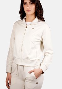 Giacca cream con zip in morbido tessuto, dotata di colletto alto, polsini a costine e dettagli in oro. Abbinata a pantaloni coordinati.