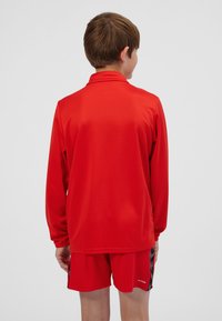 Maglietta sportiva rossa a maniche lunghe con collo alto, tessuto leggero e vestibilità comoda, abbinata a pantaloncini sportivi rossi coordinati.