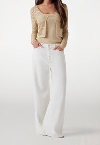 Beige neuletakki beigeä toppia vasten, yhdistetty leveälahkeisiin valkoisiin farkkuihin, joissa on etutaskut ja nappisuljin.