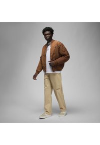 Giubbotto bomber trapuntato marrone con logo nero, t-shirt bianca, pantaloni beige e sneakers chiare, caratterizzato da una vestibilità rilassata e un design casual.