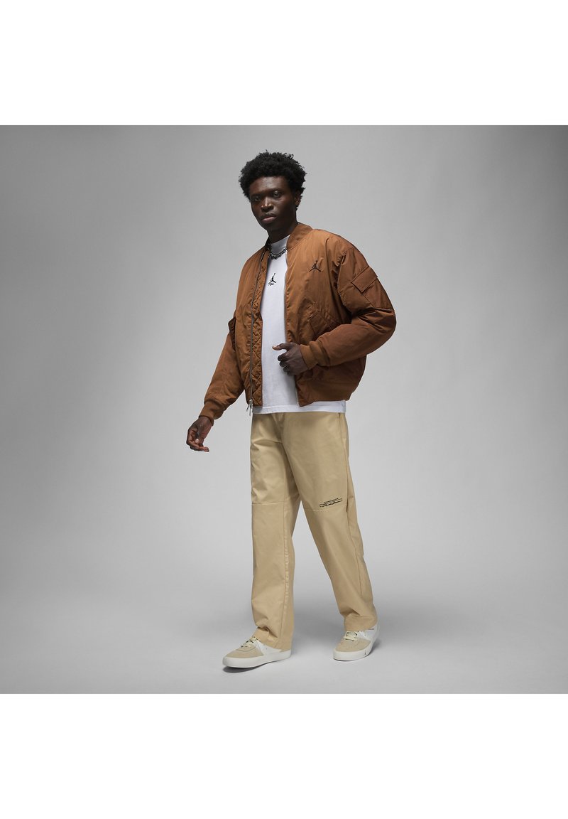 Braune gesteppte Bomberjacke mit schwarzem Logo, weißes T-Shirt, beige Hose und hellfarbene Sneakers, mit einem entspannten Schnitt und lässigem Design.