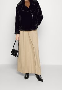 Sort jakke i imiteret pels med lynlåslukning, tekstureret overflade og bred krave, kombineret med beige plisserede bukser og sorte støvler med hæl.