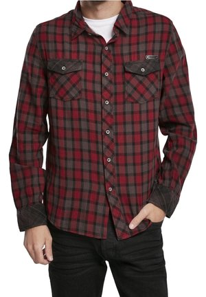 Mann trägt ein rot-schwarz kariertes Hemd über einem weißen T-Shirt, mit einer Hand in der Tasche und der anderen hängend.