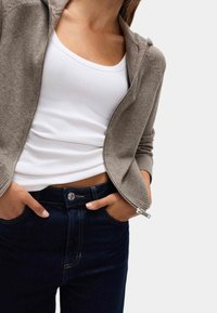 Krémová zipová mikina s pružnými manžetami, položená na bílé tílko, spárovaná s tmavě modrými slim kalhotami s kovovým knoflíkem a šitím.