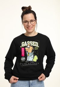 Svart sweatshirt med en tecknad grafik som föreställer en karaktär i labbrock, gul slips och texten "GASSED" i fet grön och rosa.