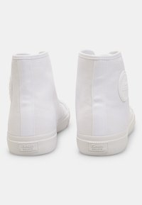 Tenisi înalți din pânză albă cu o suprafață texturată, patch rotund cu logo pe lateral și talpă din cauciuc alb. Vederea din spate evidențiază designul minimalist.