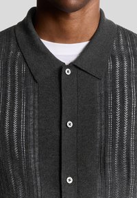Diepgrijze gebreide polo met een knoopsluiting, voorzien van een kraag en getextureerde verticale strepen. De knopen hebben een glanzende afwerking.
