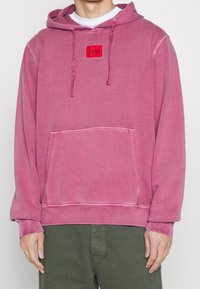Różowa bluza z kapturem ze sznurkiem, kieszenią z przodu, ściągaczami na rękawach i dole, z czerwonym prostokątnym naszywką z logo. Miękka, szczotkowana faktura tkaniny.
