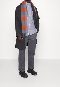 Tummanharmaa pitkä takki, sininen paita, harmaat housut ja mustat kumipohjaiset saappaat. Raidallinen oranssi ja harmaa huivi kaulan ympärillä.