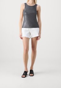 Kvinna klädd i en ärmlös glänsande mörkgrå topp, vita fransade denimshorts och svarta öppen-toe klackskor som står på en enkel bakgrund.