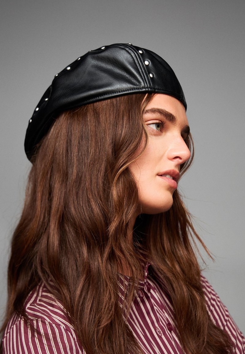 Černá kožená čepice se cvočkovými akcenty, kulatého tvaru, hladké textury. Hnědé vlnité vlasy částečně viditelné. Model nosí pruhovanou košili.