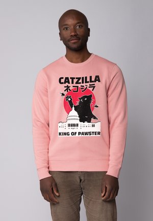 Sweat-shirt rose clair avec un graphique central représentant un chat noir sur un fond rouge, texte en gras lisant "CATZILLA ROI DES PAWS".