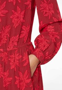 Abito floreale rosso con maniche lunghe, polsini arricciati e cucitura in vita. Caratterizzato da un tessuto leggero e testurizzato con motivi di fiori rosa.